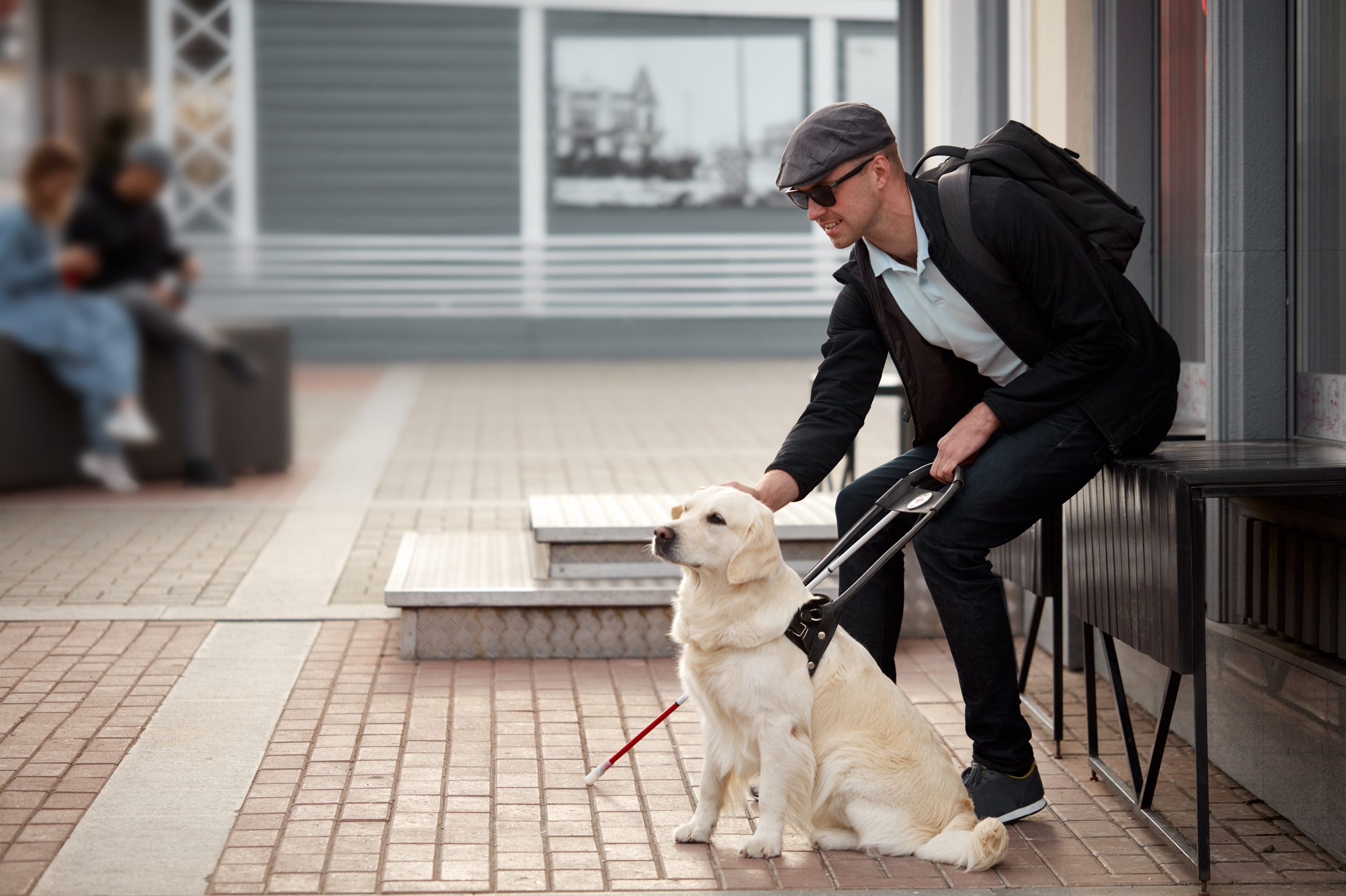 Blind man on patio petting his guide dog and holding a white cane